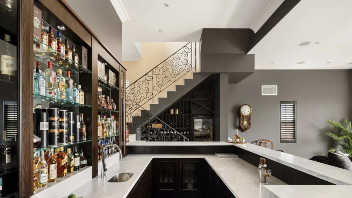 Home bar with a white stone benchtop, glass shelving stocked with spirits and a wrought-iron staircase.